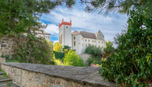 Die historische weiße Ortenburg in Bautzen mit ihrem rot gedeckten Turm und ihrer kunstvollen Architektur steht inmitten von üppigem Grün und Steinmauern, die an einem hellen, teilweise bewölkten Tag durch die Äste der Bäume zu sehen sind.