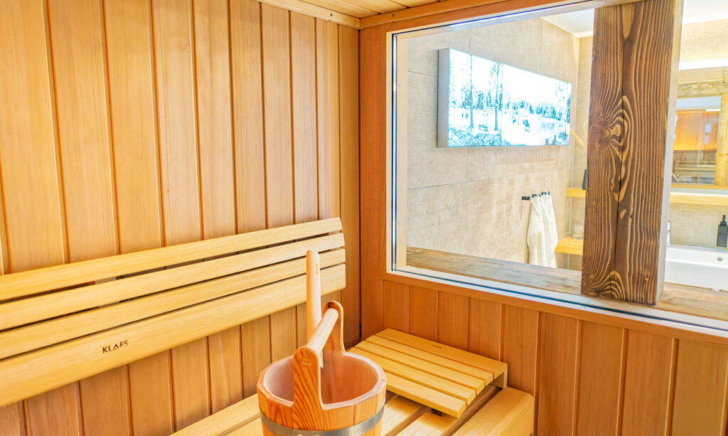 Eine moderne Holzsauna mit Bänken, einem hölzernen Eimer und einer Schöpfkelle sowie einem großen Fenster, das in ein helles Badezimmer mit Handtüchern und einem an der Wand montierten Fernseher führt.