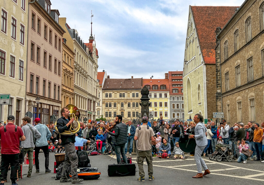 Eine lebhafte Straßenvorstellung zieht eine große Menschenmenge auf einem europäischen Stadtplatz an, wo Musiker auf Blechblasinstrumenten spielen und die Menschen, umgeben von historischen Gebäuden, unter einem teilweise bewölkten Himmel zusehen.