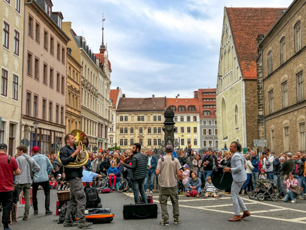 Eine lebhafte Straßenvorstellung zieht eine große Menschenmenge auf einem europäischen Stadtplatz an, wo Musiker auf Blechblasinstrumenten spielen und die Menschen, umgeben von historischen Gebäuden, unter einem teilweise bewölkten Himmel zusehen.