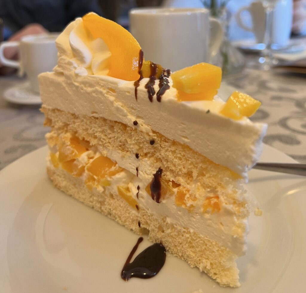 Ein Stück Schichtkuchen mit Sahne, Fruchtstückchen und Schokostreusel liegt auf einem Teller - perfekt für einen Nachmittag in einem der schönen Cafés in der Oberlausitz. Im Hintergrund: Tassen, Gläser, eine gelbe Blume in einer Vase und Menschen an einem Tisch.