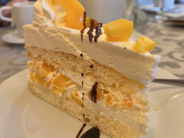 Ein Stück Schichtkuchen mit Sahne, Fruchtstückchen und Schokostreusel liegt auf einem Teller - perfekt für einen Nachmittag in einem der schönen Cafés in der Oberlausitz. Im Hintergrund: Tassen, Gläser, eine gelbe Blume in einer Vase und Menschen an einem Tisch.