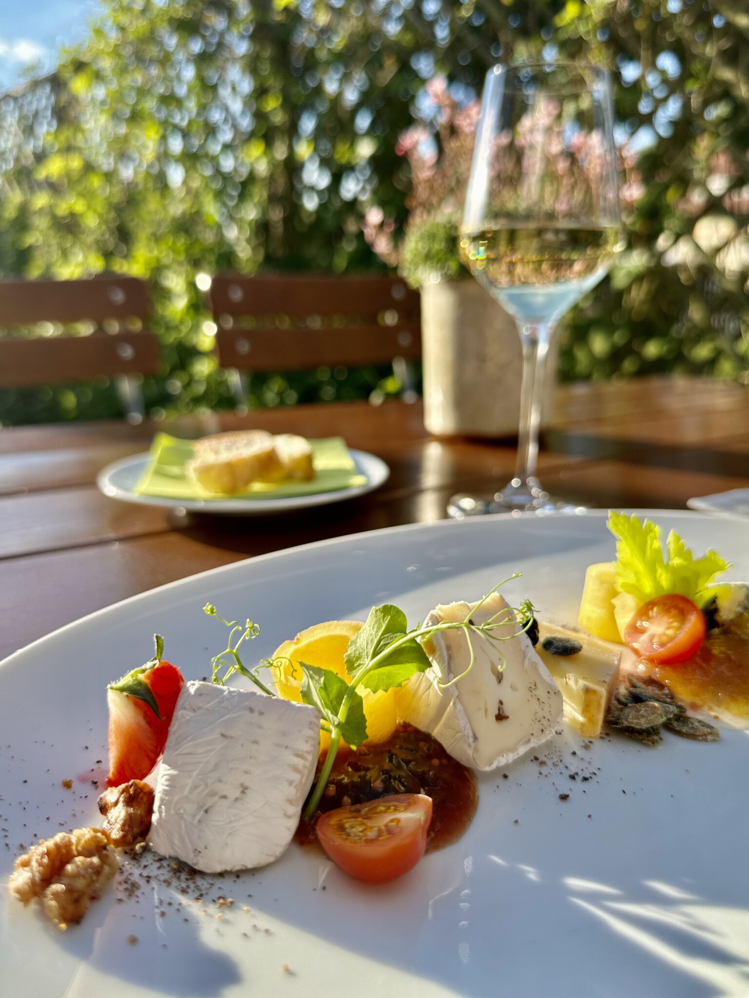 Eine Nahaufnahme eines eleganten Käsetellers in Lindners Grenzschänke mit frischen Tomaten, Nüssen und Beilagen steht auf einem Tisch im Freien, mit Sonnenlicht, einem Glas Weißwein und Brot im Hintergrund inmitten von grünem Laub.