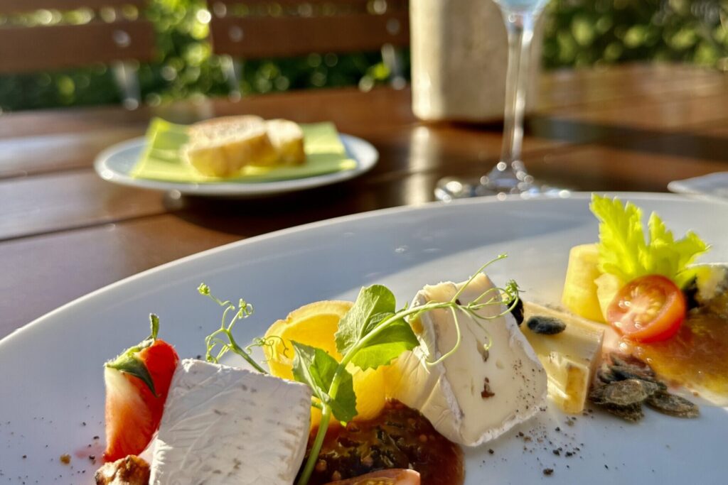 Eine Nahaufnahme eines eleganten Käsetellers in Lindners Grenzschänke mit frischen Tomaten, Nüssen und Beilagen steht auf einem Tisch im Freien, mit Sonnenlicht, einem Glas Weißwein und Brot im Hintergrund inmitten von grünem Laub.