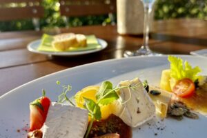 Eine Nahaufnahme eines eleganten Käsetellers in Lindners Grenzschänke mit frischen Tomaten, Nüssen und Beilagen steht auf einem Tisch im Freien, mit Sonnenlicht, einem Glas Weißwein und Brot im Hintergrund inmitten von grünem Laub.