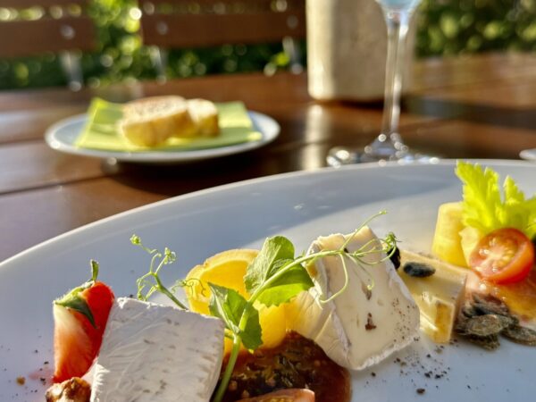 Eine Nahaufnahme eines eleganten Käsetellers in Lindners Grenzschänke mit frischen Tomaten, Nüssen und Beilagen steht auf einem Tisch im Freien, mit Sonnenlicht, einem Glas Weißwein und Brot im Hintergrund inmitten von grünem Laub.