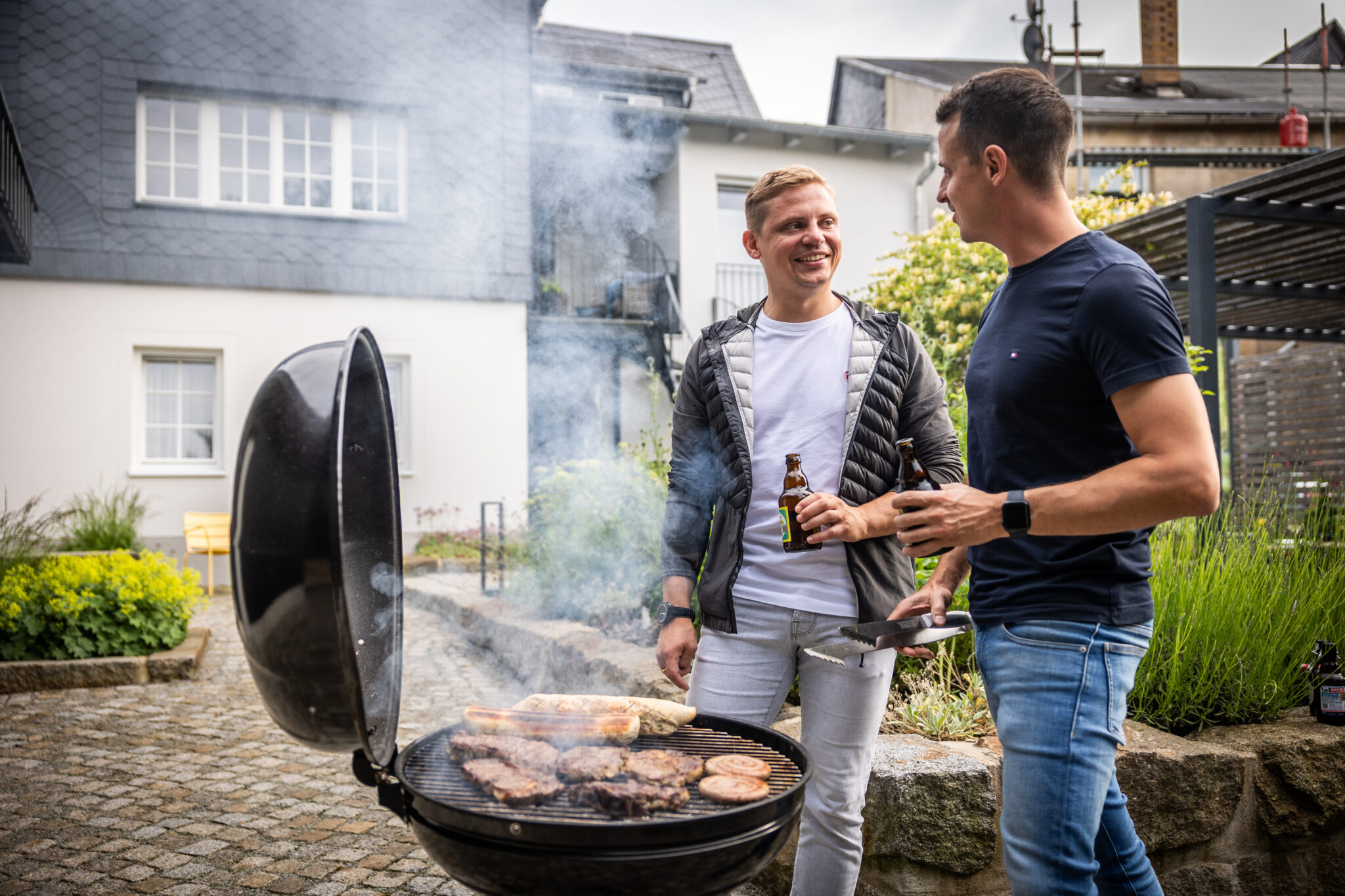 Zwei Männer stehen in einem Hinterhof eines Ferienhauses für Gruppen an einem rauchenden Grill, unterhalten sich und lächeln. Der eine hält ein Getränk in der Hand, der andere eine Grillzange, während auf dem offenen Grill gegrilltes Fleisch brutzelt. Häuser und Grünflächen rahmen die Szene ein.