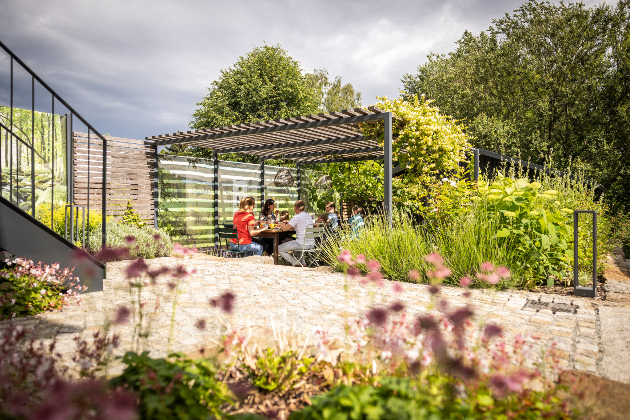 Menschen sitzen an einem Tisch unter einer modernen Pergola in einem üppigen Garten eines Ferienhauses für Gruppen, umgeben von Grün und blühenden Blumen, mit einem bewölkten Himmel über sich. Die Szene ist einladend und entspannt.