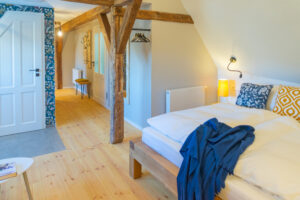 Ein gemütliches Schlafzimmer mit Holzboden, sichtbaren Balken, einem Doppelbett mit weißer Bettwäsche und gelben Kissen, einer blauen Bettdecke und einer Nachttischlampe - perfekt für eine Auszeit zu zweit in der Oberlausitz. Eine gemusterte Tapete säumt den offenen Flur.