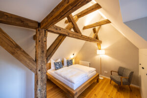 Ein gemütliches Schlafzimmer im Dachgeschoss mit freiliegenden Holzbalken, einem Doppelbett und einem gepolsterten Sessel auf poliertem Boden - perfekt für eine Auszeit zu zweit in der Oberlausitz. Warme Beleuchtung und gemusterte Kissen schaffen eine einladende Atmosphäre.