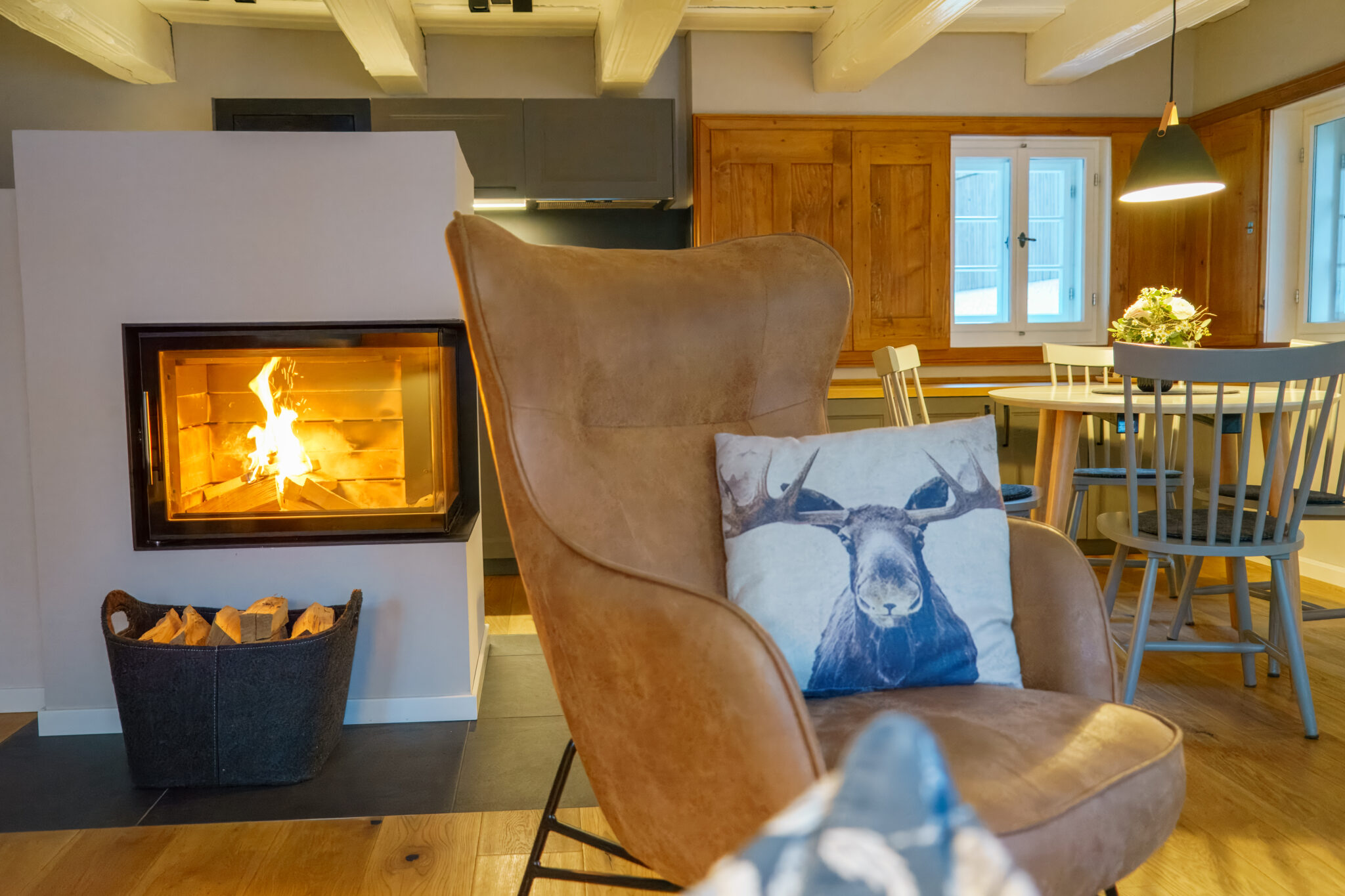 Ein gemütliches Wohnzimmer mit modernem Kamin, gestapeltem Brennholz und einem braunen Sessel mit Elchkissen - Einfach mal raus - Wohlfühlzeit im Marktquartier 5. Warme Holzakzente und sanfte Beleuchtung schaffen eine einladende Atmosphäre am Esstisch am Fenster.