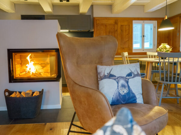 Ein gemütliches Wohnzimmer mit modernem Kamin, gestapeltem Brennholz und einem braunen Sessel mit Elchkissen - Einfach mal raus - Wohlfühlzeit im Marktquartier 5. Warme Holzakzente und sanfte Beleuchtung schaffen eine einladende Atmosphäre am Esstisch am Fenster.
