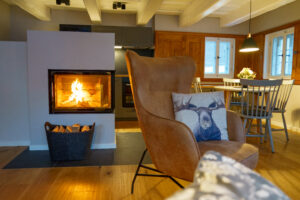 Ein gemütliches Wohnzimmer mit einem modernen Kamin, einem Ledersessel mit Elchkissen, einem Korb mit Brennholz und einem Esstisch mit Stühlen am Fenster. Die warme Beleuchtung schafft eine einladende Atmosphäre im Marktquartier 5 - Ferienapartments in der Oberlausitz.