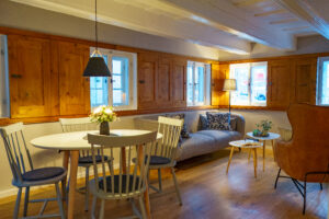 Ein gemütliches Wohnzimmer mit einem hellen Holzboden, einem runden Esstisch mit vier Stühlen, einem grauen Sofa mit gemusterten Kissen, einem Sessel, kleinen Tischen mit Lampen, hölzernen Wandpaneelen und mehreren Fenstern, die natürliches Licht hereinlassen. Perfekter Urlaub im Marktquartier 5 - Ferienapartments in der Oberlausitz