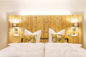 Ein gemütliches Doppelbett mit weißer Bettwäsche und blattgemusterten Kissen lädt zum Entspannen ein - Einfach mal raus - Wohlfühlzeit im Marktquartier 5 - an einem hölzernen Kopfteil mit Wandlampen an beiden Seiten.