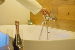 Eine Person in weißem Bademantel dreht den Wasserhahn einer Badewanne auf, das Wasser fließt. Im Vordergrund laden Champagner und zwei Gläser zur Entspannung ein - Einfach mal raus - Wohlfühlzeit im Marktquartier 5 verspricht Erholung und Luxus pur.