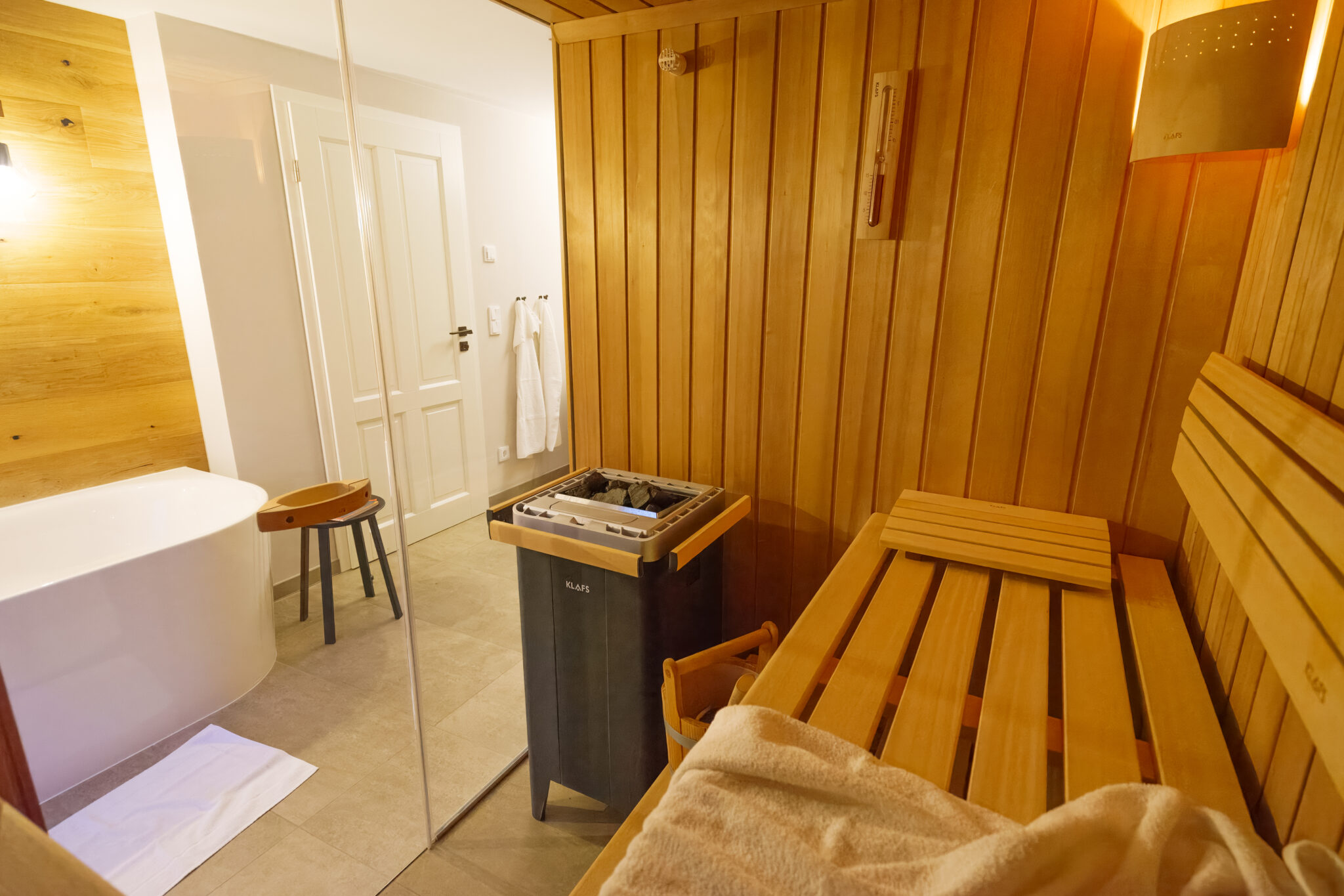 Ein moderner Saunaraum mit Holzbänken, einem Ofen und einem Handtuch lädt zum Entspannen ein. Genießen Sie die Wohlfühlzeit im Marktquartier 5 mit einem Badezimmer, das mit einer weißen Badewanne, einem Hocker und Bademänteln ausgestattet ist. Der Raum ist sauber und gut beleuchtet.