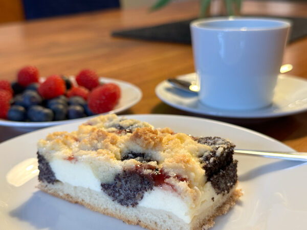 Nahaufnahme eines Stücks Oberlausitzer Kleckskuchen mit Mohn-, Quark- und Marmeladenschicht - eine der kulinarischen Spezialitäten der Oberlausitz - auf einem weißen Teller. Im Hintergrund stehen eine Tasse Kaffee und ein Teller mit Blaubeeren und Himbeeren.