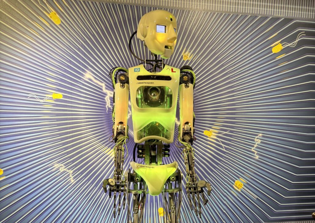 Ein humanoider Roboter mit durchsichtigem Brustkorb und neongrünen Teilen steht auf einem Podest, das vor einer grafischen Kulisse aufgebaut ist - ein auffälliges Highlight, das perfekt zu den Schlechtwettertipps Oberlausitz mit Teenagern passt.