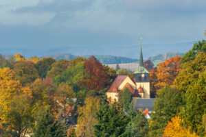 Die Dorfkirche in Neusalza-Spremberg mit einem hohen Kirchturm steht zwischen bunten Herbstbäumen, mit sanften Hügeln und einem bewölkten blauen Himmel im Hintergrund.