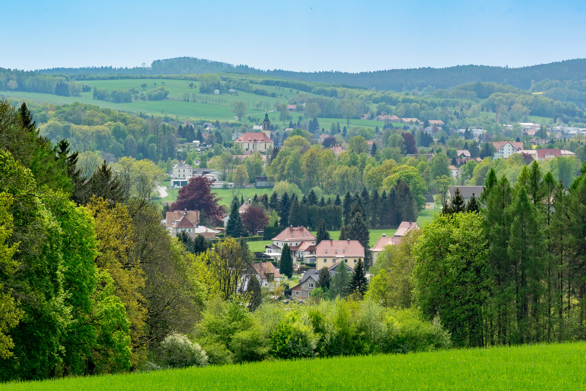 Eine malerische Landschaft mit einem kleinen Dorf mit verstreuten Häusern, umgeben von üppig grünen Bäumen und sanften Hügeln unter einem klaren, blauen Himmel.
