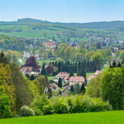 Eine malerische Landschaft mit einem kleinen Dorf mit verstreuten Häusern, umgeben von üppig grünen Bäumen und sanften Hügeln unter einem klaren, blauen Himmel.