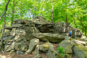 Große, geschichtete Felsformation, umgeben von grünen Bäumen in einem Wald am Oberlausitzer Bergweg, mit einer blau gekleideten Person, die auf den Felsen rechts sitzt und das Wandern ohne Gepäck und die Natur an einem sonnigen Tag genießt.