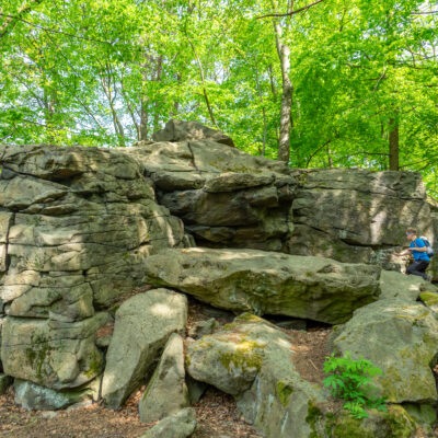 Große, geschichtete Felsformation, umgeben von grünen Bäumen in einem Wald am Oberlausitzer Bergweg, mit einer blau gekleideten Person, die auf den Felsen rechts sitzt und das Wandern ohne Gepäck und die Natur an einem sonnigen Tag genießt.