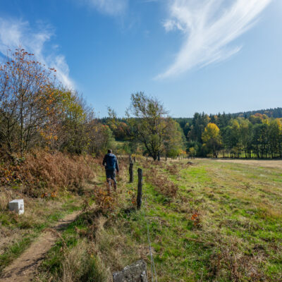 Eine Person genießt das Wandern ohne Gepäck und spaziert auf einem schmalen Feldweg durch grasbewachsene Felder und herbstliche Bäume unter einem strahlend blauen Himmel. Im Hintergrund erheben sich bewaldete Hügel, die Teil des Oberlausitzer Bergwegs sind.
