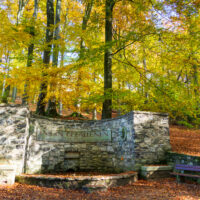 Ein steinerner Brunnen mit einer Aufschrift steht zwischen hohen Bäumen mit gelben Herbstblättern; herabgefallene Blätter bedecken den Boden, und in der Nähe stehen Holzbänke. Herbstidyll am Oberlausitzer Bergweg.