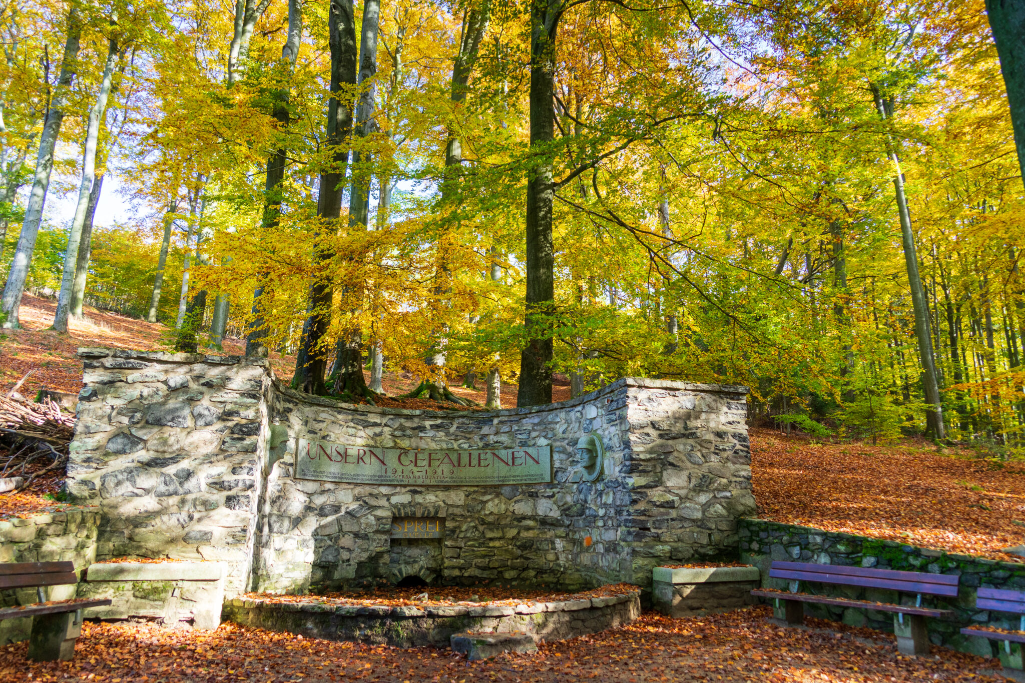 Ein steinerner Brunnen mit einer Aufschrift steht zwischen hohen Bäumen mit gelben Herbstblättern; herabgefallene Blätter bedecken den Boden, und in der Nähe stehen Holzbänke. Herbstidyll am Oberlausitzer Bergweg.