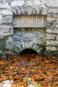 Ein steinernes Bauwerk mit bogenförmiger Öffnung, darüber in goldenen Lettern die Inschrift SPREE QUELLE. An der Basis schwimmen Herbstblätter auf dem Wasser, die auf die Quelle der Spree am Kottmar direkt am Oberlausitzer Bergweg hinweisen.