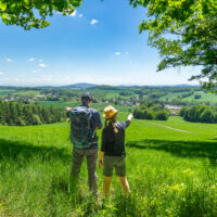 Zwei Menschen mit Hüten und Rucksäcken stehen auf einer grasbewachsenen Anhöhe am Oberlausitzer Bergweg. Der eine zeigt in Richtung eines fernen Dorfes, das von grünen Feldern und Hügeln umgeben ist - perfekt für Wandern ohne Gepäck unter strahlend blauem Himmel.