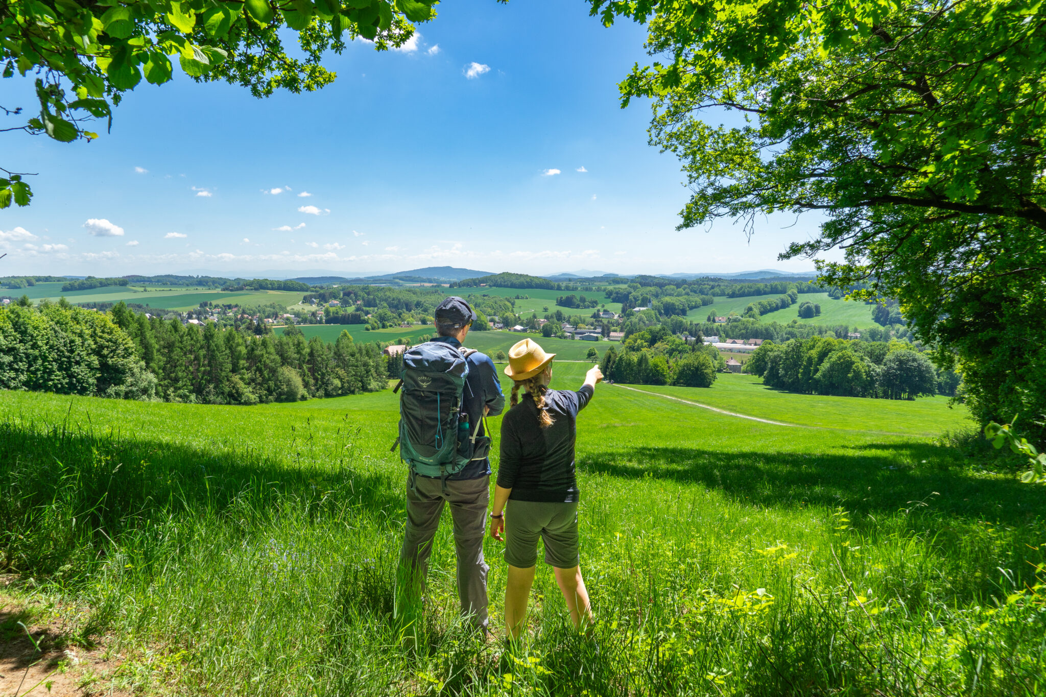 Zwei Menschen mit Hüten und Rucksäcken stehen auf einer grasbewachsenen Anhöhe am Oberlausitzer Bergweg. Der eine zeigt in Richtung eines fernen Dorfes, das von grünen Feldern und Hügeln umgeben ist - perfekt für Wandern ohne Gepäck unter strahlend blauem Himmel.