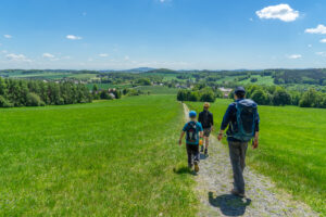 Drei Personen, zwei Erwachsene und ein Kind, wandern auf einem Kiesweg durch eine grüne Wiese unter strahlend blauem Himmel in der Nähe der Ferienwohnungen Oberlausitz, in der Ferne sind Bäume und ein Dorf zu sehen - perfekt für Eure Auszeit im Marktquartier 5.
