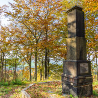 Auf einer sonnenbeschienenen Waldlichtung auf dem Kottmar steht ein hohes Steinmonument, umgeben von herbstlichen Bäumen mit goldenen und orangefarbenen Blättern. Der Himmel ist klar und blau, in der Ferne sind Hügel zu sehen - ein perfekter Ort für Wandern ohne Gepäck.