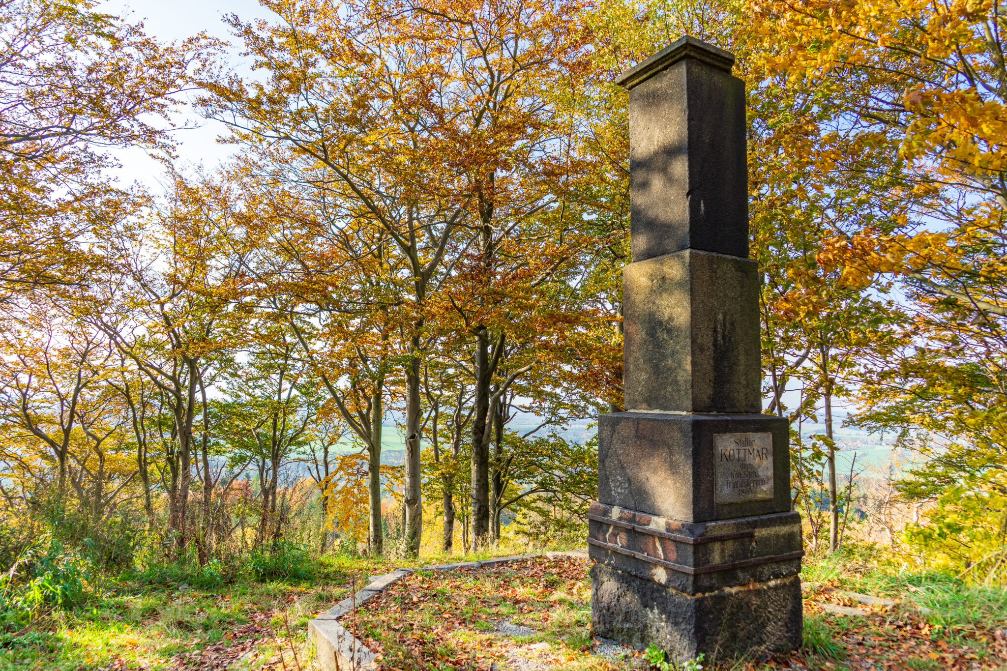 Auf einer sonnenbeschienenen Waldlichtung auf dem Kottmar steht ein hohes Steinmonument, umgeben von herbstlichen Bäumen mit goldenen und orangefarbenen Blättern. Der Himmel ist klar und blau, in der Ferne sind Hügel zu sehen - ein perfekter Ort für Wandern ohne Gepäck.