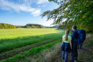 Zwei Wanderer mit Rucksäcken stehen am Rande einer grünen Wiese und bewundern die Aussicht auf den Oberlausitzer Bergweg ohne Gepäck, umgeben von üppigem Laub unter einem blauen Himmel mit vereinzelten Wolken.
