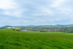 In der Ferne erstreckt sich ein weites, grünes Feld mit einem kleinen Dorf zwischen Bäumen und sanften Hügeln unter einem bewölkten Himmel. Am Horizont sind gelbe Flecken von Feldfrüchten und ferne Berge zu sehen.