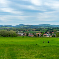 Zwei Personen gehen über eine weite, grüne Wiese in Richtung eines kleinen Dorfes und genießen den Oberlausitzer Bergweg und Wandern ohne Gepäck, mit Häusern und Bäumen vor sanften Hügeln und einem bewölkten Himmel in der Ferne.