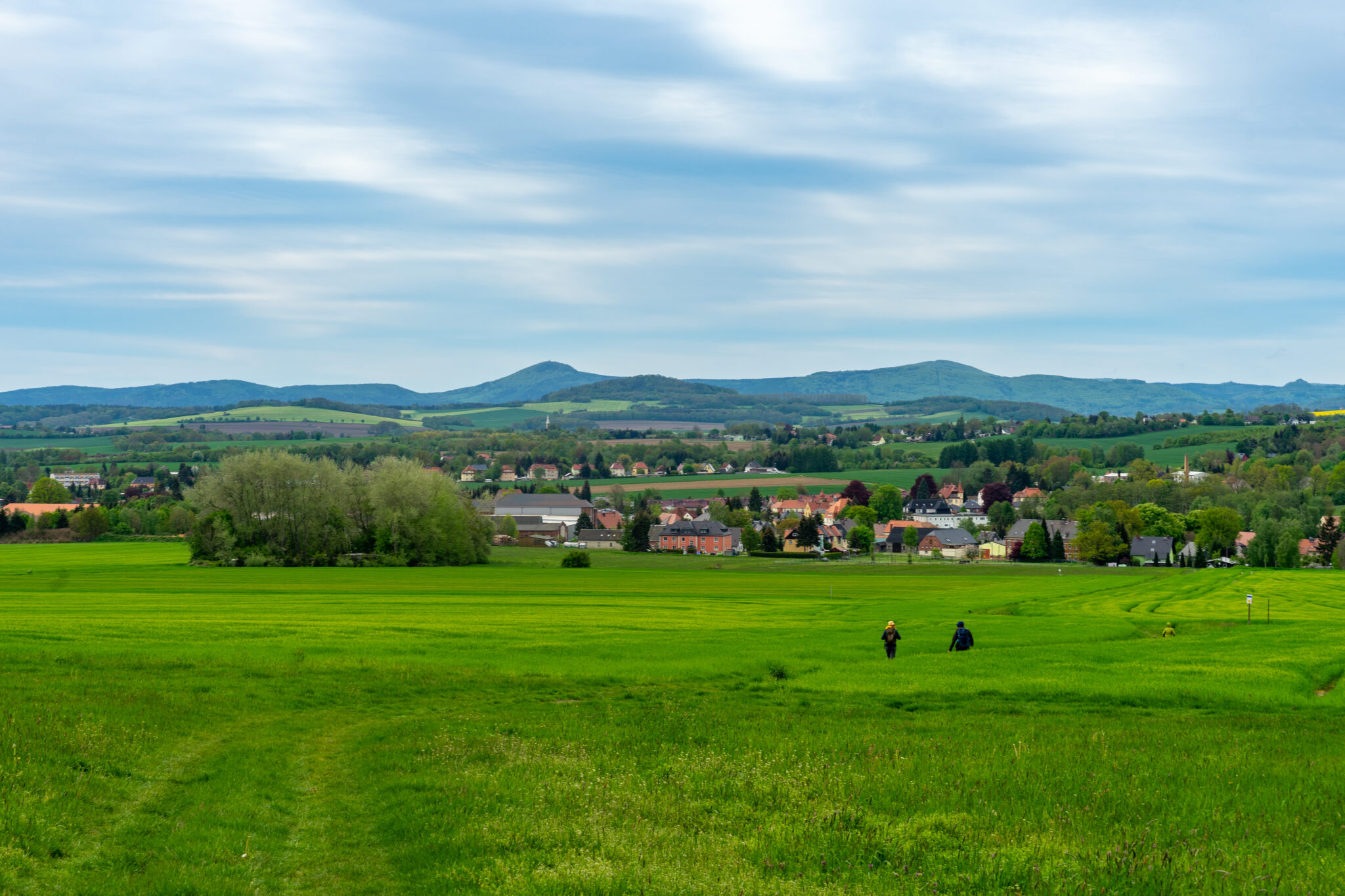 Zwei Personen gehen über eine weite, grüne Wiese in Richtung eines kleinen Dorfes und genießen den Oberlausitzer Bergweg und Wandern ohne Gepäck, mit Häusern und Bäumen vor sanften Hügeln und einem bewölkten Himmel in der Ferne.