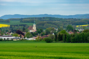 Blick auf Eibau, ein ländliches Dorf in der Oberlaustiz, mit einer Kirche mit grüner Kuppel, umgeben von Bäumen, grünen Grasfeldern und gelben Feldfrüchten, mit sanften Hügeln und Wald im Hintergrund unter einem leicht bewölkten Himmel.