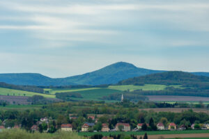 Ein kleines Dorf mit verstreuten Häusern und einem Kirchturm liegt inmitten grüner Felder und Bäume in der Oberlausitz. Im Hintergrund erheben sich sanfte Hügel und die bewaldeten Berge der Lausche unter einem bewölkten Himmel. Auf dem Oberlausitzer Bergweg, Wandern ohne Gepäck vom Marktquartier 5 aus gut erlebbar.