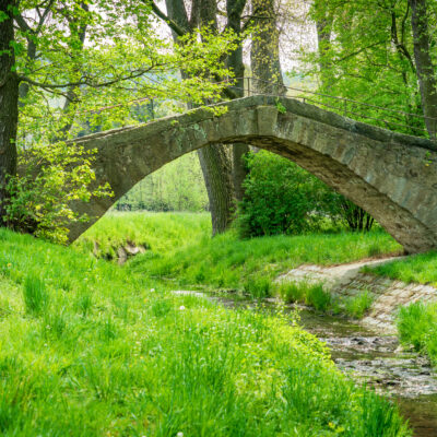 Die Himmelsbrücke in Sohland, eine alte Steinbogenbrücke, überquert die Spree in einem üppig grünen Park am Oberlausitzer Bergweg - perfekt für Wanderungen ohne Gepäck zwischen frischem Frühlingslaub und Wildblumen.