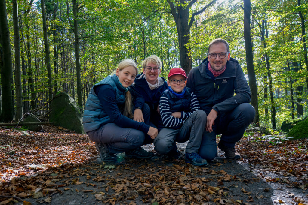 Vier Personen, zwei Erwachsene und zwei Kinder, knien und lächeln gemeinsam auf einem mit Blättern bedeckten Waldweg. Das Sonnenlicht fällt durch die Bäume im Hintergrund und schafft eine fröhliche Atmosphäre im Freien. Wandern auf dem Oberlausitzer Bergweg.