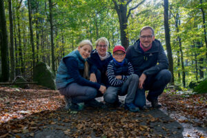 Vier Personen, zwei Erwachsene und zwei Kinder, knien und lächeln gemeinsam auf einem mit Blättern bedeckten Waldweg. Das Sonnenlicht fällt durch die Bäume im Hintergrund und schafft eine fröhliche Atmosphäre im Freien. Wandern auf dem Oberlausitzer Bergweg.