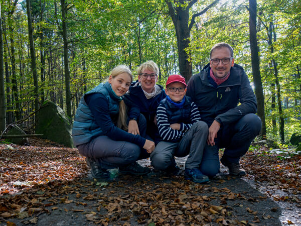 Vier Personen, zwei Erwachsene und zwei Kinder, knien und lächeln gemeinsam auf einem mit Blättern bedeckten Waldweg. Das Sonnenlicht fällt durch die Bäume im Hintergrund und schafft eine fröhliche Atmosphäre im Freien. Wandern auf dem Oberlausitzer Bergweg.