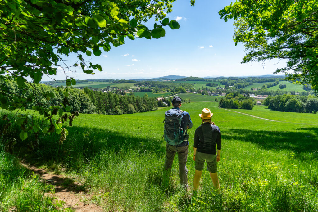 Zwei Menschen mit Rucksäcken stehen auf einer grasbewachsenen Bergkuppe am Oberlausitzer Bergweg und blicken auf eine grüne, ländliche Landschaft mit Feldern, Bäumen und fernen Hügeln unter einem strahlend blauen Himmel. Wandern ohne Gepäck macht die Reise leicht und unbeschwert. Baumzweige rahmen die Szene ein.