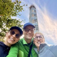 Wandernde vor dem neuen Aussichtsturm Jüttelsberg (Jitrovník) bei Schluckenau. Sie lächeln auf einem Selfie im Freien, im Hintergrund ist ein hoher, spiralförmiger Aussichtsturm auf dem Jüttelsberg (Jitrovník) zu sehen.