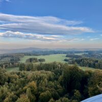 Blick aus großer Höhe auf eine weitläufige, hügelige Waldlandschaft unter blauem Himmel mit vereinzelten Wolken.